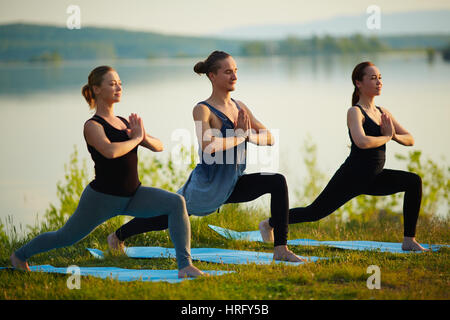 Group of three young people practicing yoga on river bank, stretching hips and holding hands in namaste Stock Photo