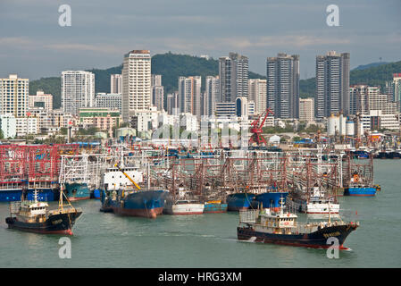 A view of Sanya harbour in China with fishing boats and ships moored and the skyscrapers of the city behind with a ship boat leaving harbour. Stock Photo