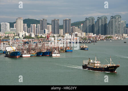A view of Sanya harbour in China with fishing boats and ships moored and the skyscrapers of the city behind with a ship boat leaving harbour. Stock Photo