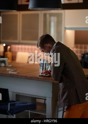 Side view of man standing in dim modern kitchen leaning on isle table and reading messages in smartphone Stock Photo