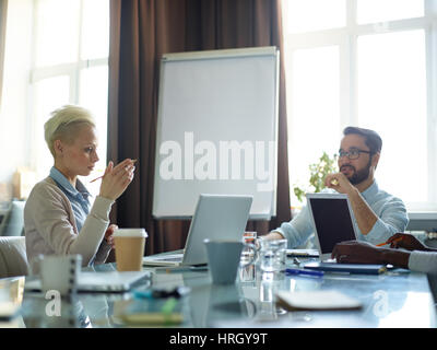 Creative business people having long discussion over work related issues thinking and sharing ideas while sitting at messy meeting table in modern con Stock Photo