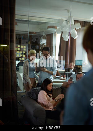 Modern office room made in warm brown tones filled with creative business people busy discussing plans and working at computers, shot from behind glas Stock Photo