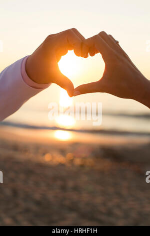 Sonnenaufgang am Strand, Paar formt Herz mit Haenden - sunset at the beach, couple makes a heart with hands Stock Photo