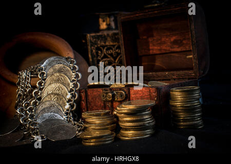 Treasure chest, pile and pillar of coins, a ceramic bowl filled with jewelry coins and candle lamp in dark environment Stock Photo