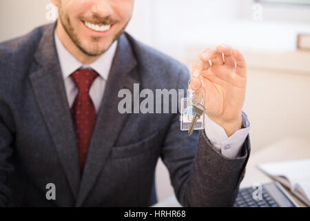 Cropped toned image of handsome middle aged realtor in classic suit holding key from new apartment to his clients and smiling while sitting in office. Stock Photo