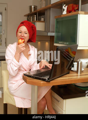 Junge Frau im Morgenmantel arbeitet am Notebook und isst einen Apfel - young woman in bath robe using laptop and eating an apple Stock Photo