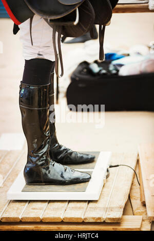 Rider wearing shiny black riding boots at the weigh in  on weighing scale, holding a saddle, before or after a race. Stock Photo