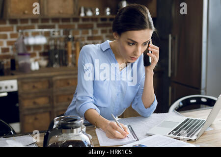 Concentrated young woman using laptop and smartphone while making notes Stock Photo