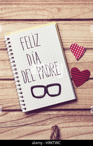 the text feliz dia del padre, happy fathers day in spanish written in the page of a notebook, a pair of black eyeglasses and some red hearts on a rust Stock Photo