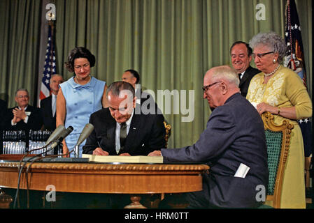 PRESIDENT LYNDON B. JOHNSON signs the Medicare Bill on 30 July 1965 watched by his wife Lady Bird in blue and former President Harry S.Truman seated right with his wife Bess in yellow. Vice President Hubert Humphrey stands at right. Photo: White House official Stock Photo