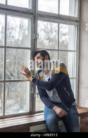 Emotional sad bearded man looking through the window. He is looking worried, depressed, thoughtful and lonely. waiting concept. Stock Photo