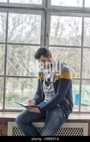 Portrait of a casual bearded hipster man writing and making notes in copybook or notepad at home. Big windows backdround. handwriting Stock Photo