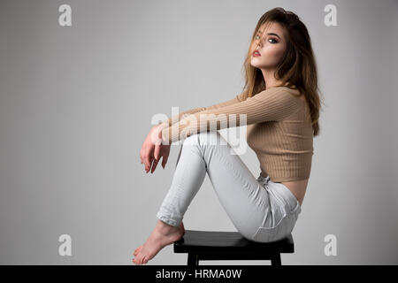 Portrait of young woman wearing jeans and biege sweater sitting on a stool Stock Photo