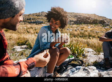 Happy young african woman eating sandwich and smiling while relaxing with friends during hike. Group of hikers taking break during country hike. Stock Photo