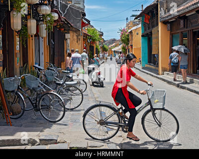 Young Vietnamese woman in red ao dai dress riding bicycle. Hoi An Ancient Town, Quang Nam Province, Vietnam. Stock Photo