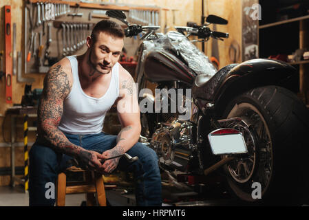 It is my bike. Serious muscular male sitting on the chair near big wheel holding spanner while posing in the garage Stock Photo