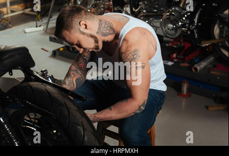 Be attentive. Young muscular man stooping down to his bike sitting in the workshop while working with it Stock Photo