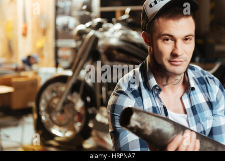 That is good. Close up of handsome man wearing stylish cap and blue checked shirt holding old exhaust tube on the foreground, smiling at camera Stock Photo