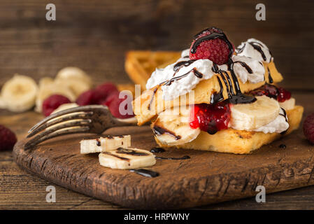 Belgian waffles with raspberries, bananas and double cream on rustic table. Stock Photo