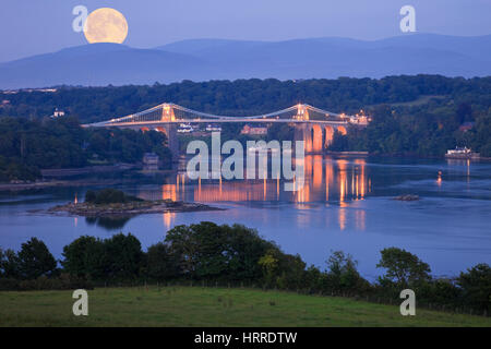 Moonrise full moon rising over Menai Strait coast with Menai suspension bridge illuminated at night. Menai Bridge, Isle of Anglesey, North Wales, UK Stock Photo