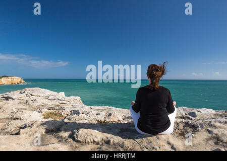 WOMAN MEDITATING ON CLIFF TOP IN YOGA POSITION FACING SEA CABO ROJO PUERTO RICO Stock Photo