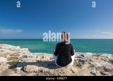 WOMAN MEDITATING ON CLIFF TOP IN YOGA POSITION FACING SEA CABO ROJO PUERTO RICO Stock Photo