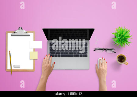 Woman work on modern laptop. Top view with clipboard, business card, glasses, plant and coffee. Pink desk in background.
