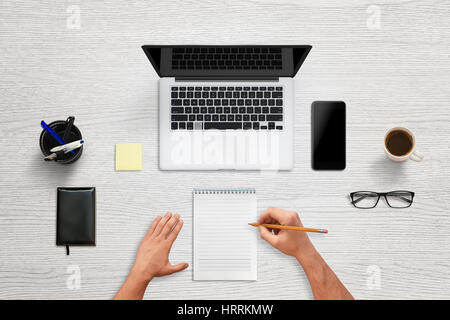 Work in office. Top view of desk with laptop computer and mobile phone. Man write on pad. Clean, blank office supplies for mockup. Stock Photo