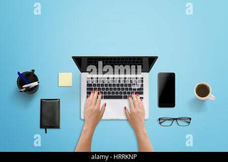 Blue work desk in modern office. Woman writing on the laptop computer. Smart phone with blank screen for mockup beside. Stock Photo