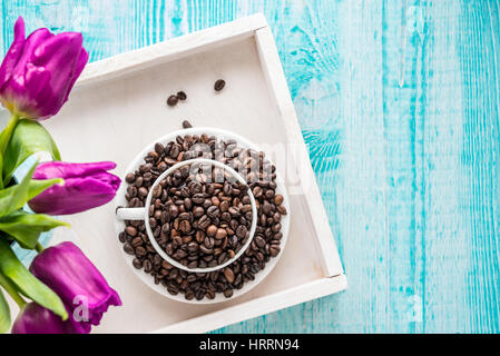 Vintage wooden tray with porcelain cup full of coffee beans and pink flowers on shabby chic mint background, top view Stock Photo