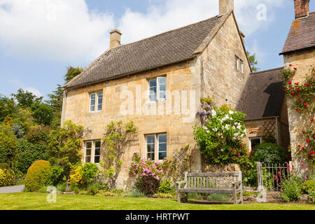 English village stone cottage with red roses, colorful flowers in the garden and wooden bench on the lawn, on a sunny, summer day . Stock Photo