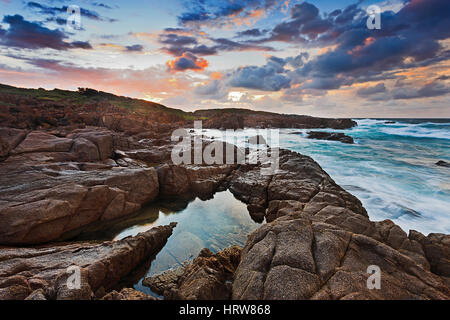 Red rugged rocks near Birubi beach of Port Stephens, NSW, Australia. Sunbeams light clouds, sea waves and water puddles between rocks. Stock Photo