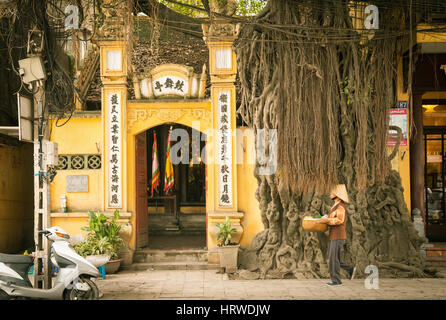 Hanoi, Vietnam - April 25, 2014: Vietnamese woman in conical hat with basket in hands is passing by the temple on the street of Old Quarter, Hanoi, Vi Stock Photo