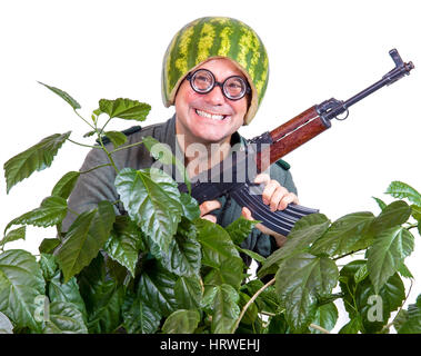 Funny cheerful soldier with a machine gun sticking out of the leaves. Crazy man in a helmet of watermelon patrolling with gun in his hands. Stock Photo