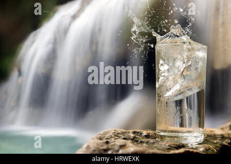 Dewy glass full of water with ice cube stands on a rock near a waterfall in the forest. A refreshing drink from nature. Ice cube falls into a glass of Stock Photo