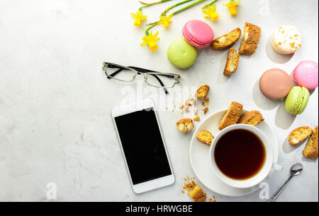 Cup of black tea with tasty almond cookies, rich in vitamins, minerals, varicolored macarons, smartphone, glasses and flowers on a white background in Stock Photo