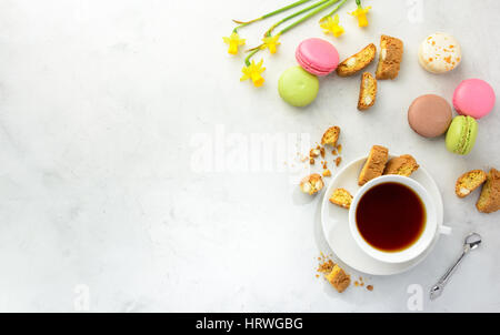 Cup of black tea with tasty almond cookies, rich in vitamins, minerals, varicolored macarons and flowers on a white background in light key. Top view  Stock Photo