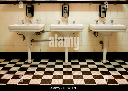 Three white porcelain sinks in an old Art Deco public bathroom with a black and white checkered tile floor. Stock Photo