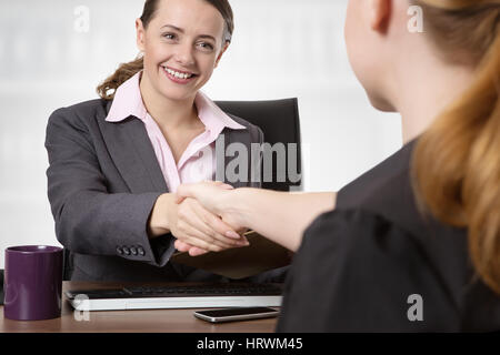 Close up office shot of two women shaking hands whilst sitting down at a desk. Stock Photo