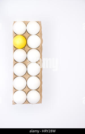 One Easter egg dyed yellow and eleven white hen's egg in a cardboard carton from above with white background and copy space. Stock Photo