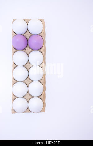 Two Easter eggs dyed lavender or lilac and ten white hen's egg in a tan cardboard carton from above with white background and copy space. Stock Photo