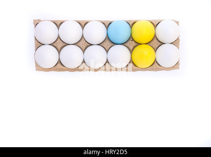 Two Easter eggs dyed yellow, one egg dyed blue and nine white hen's eggs in a tan cardboard carton from above on white background with copy space. Stock Photo