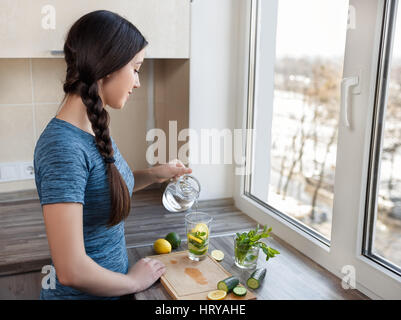 girl preparing detox water with cucumber, mint and lemon at home kitchen. Healthy drink. Weight loss dieting  concept. Process of preparing energy bev Stock Photo