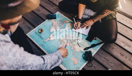 Man and woman planning tour using a world map. Woman making notes while the man is pointing at world map with travel accessories around. Stock Photo