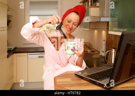 Junge Frau im Morgenmantel arbeitet am Notebook an der Kuechenbar und schenkt sich Tee ein - woman in bathrobe using laptop and drinking tea Stock Photo