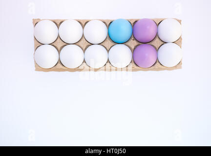Two Easter eggs dyed yellow, one egg dyed blue and nine white hen's eggs in a tan cardboard carton from above on white background with copy space. Stock Photo