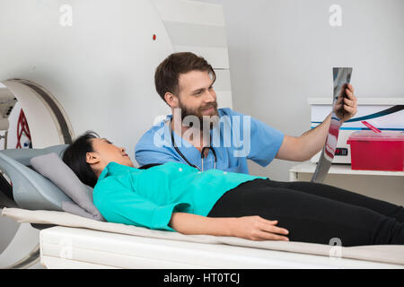 Young male radiologist showing X-ray report to female patient lying on CT scan machine at hospital Stock Photo