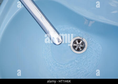 Water flows from the tap in a blue washbasin. The liquid pours out, swirling, in the open plug Stock Photo