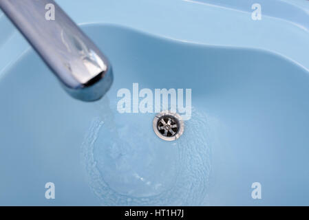 Water flows from the tap in a blue washbasin. The liquid pours out, swirling, in the open plug Stock Photo