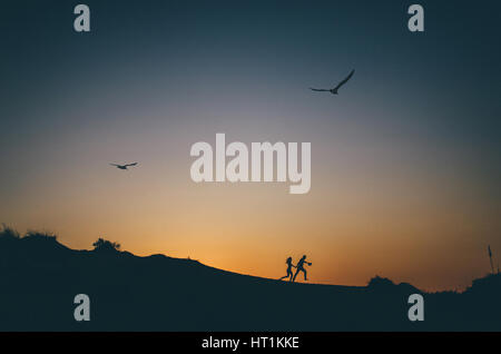 Small silhouette of happy couple holding hands and running up the hill at sunset with two seagulls flying in the sky Stock Photo